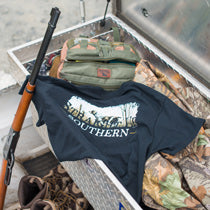 A Straight Up Southern t-shirt is draped over a box of hunting gear, including a rifle, boots, and camouflage