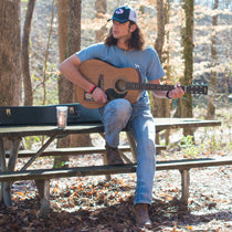 A man with long hair, a baseball cap, a guitar, and a Straight Up Southern t-shirt sits on a picnic table