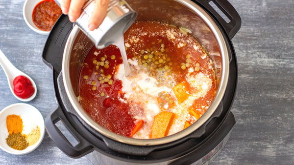 Slow cooker filled with lentils and vegetables showing a hand pouring in coconut milk