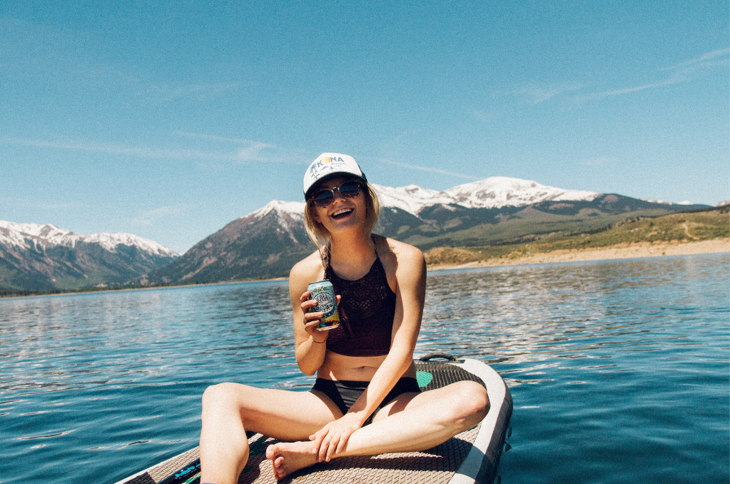 Woman sitting on a Stand up Paddleboard on the lake. She is wearing a hat and sunglasses, drinking out of a can