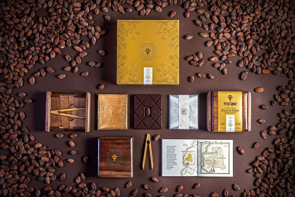 An overhead photograph of the To’ak chocolate selection, the handcrafted wooden boxes they come in, the wooden tasting utensils, and the gift set. All of them are tastefully arranged on a table, and are surrounded by cacao beans.