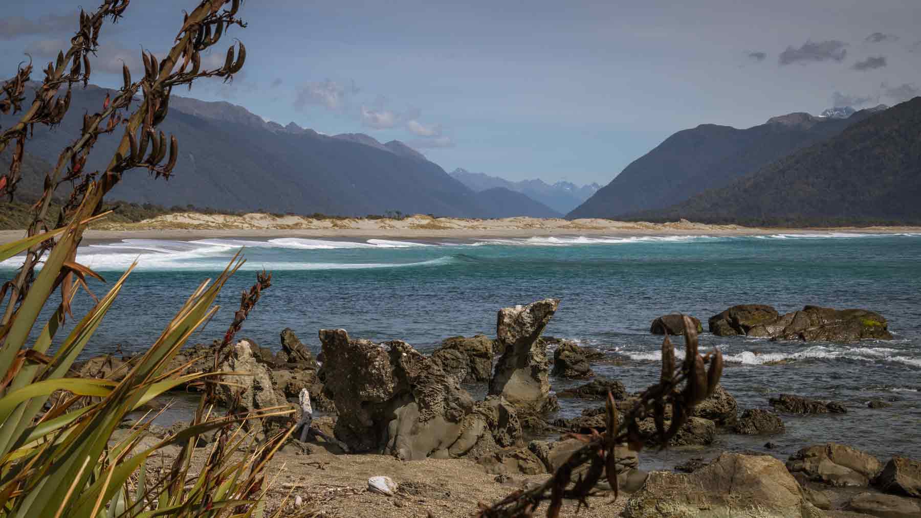View from Martins Bay, with flax, sea and sand hills in the foreground,  up the Hollyford Valley, Fiordland, New Zealand taken by Asmuss founder Fiona. Walking the Hollyford Track was a great test of the Asmuss luxury stylish sustainable travel clothing