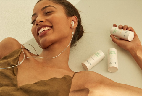 Alt text: A person in a brown tank top listens to music, holding We Are Wild skincare products.