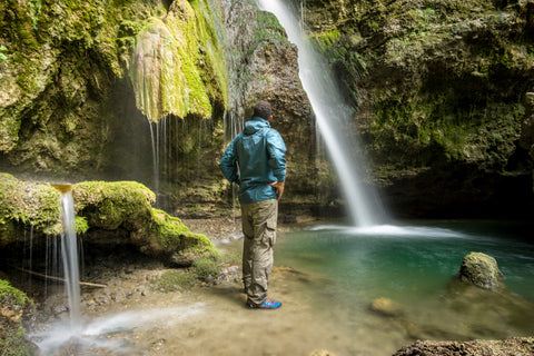 A hiker in a rain jacket stands underneath a waterfall.