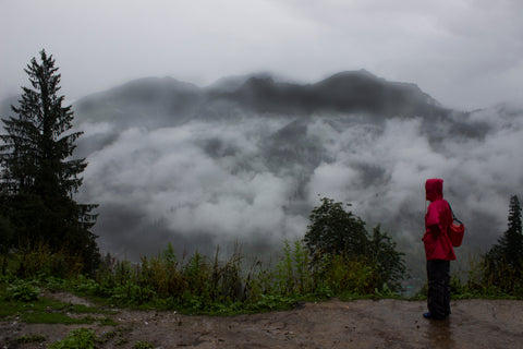 A hiker in full rain gear, including rain pants and a rain jacket stands in front of a fog-filled forest.