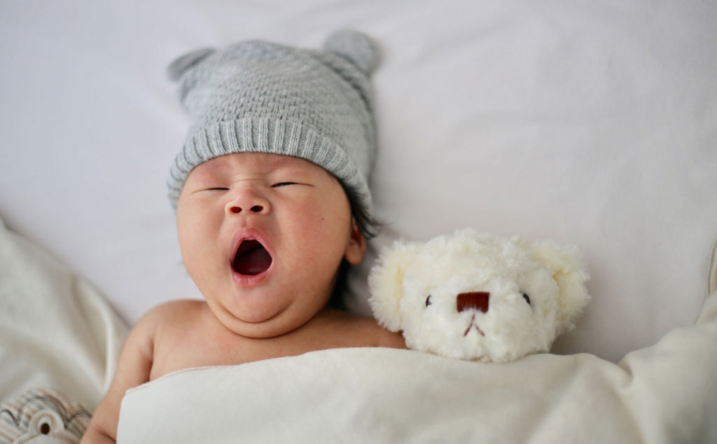 Baby in bed with his teddy. Grey woollen hat, white teddy.
