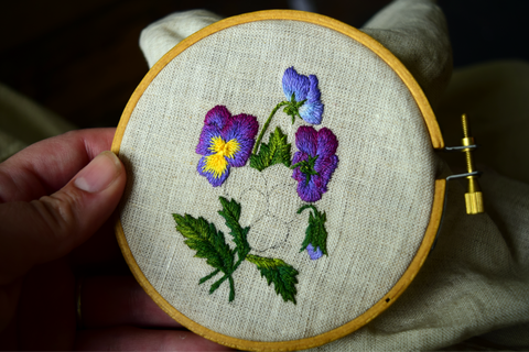 Design of purple and yellow pansies against green leaves, one flower incomplete in hoop.