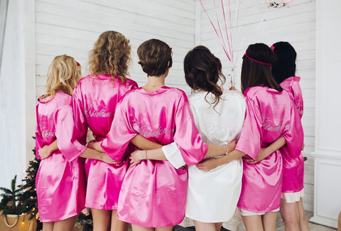 back view of woman friends who wore pink satin robes and hug the bride wore a white satin robe as an answer to the question of what is a bachelorette party