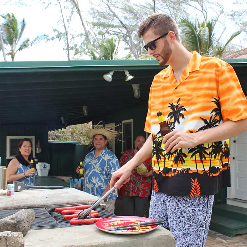 Davis Rozitis grilling hot dogs in an orange Hawaiian shirt and board shorts