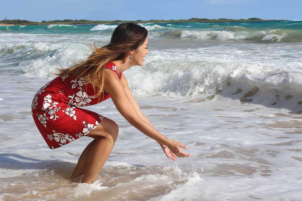 stepping into the water at the beach in a red Hawaiian dress