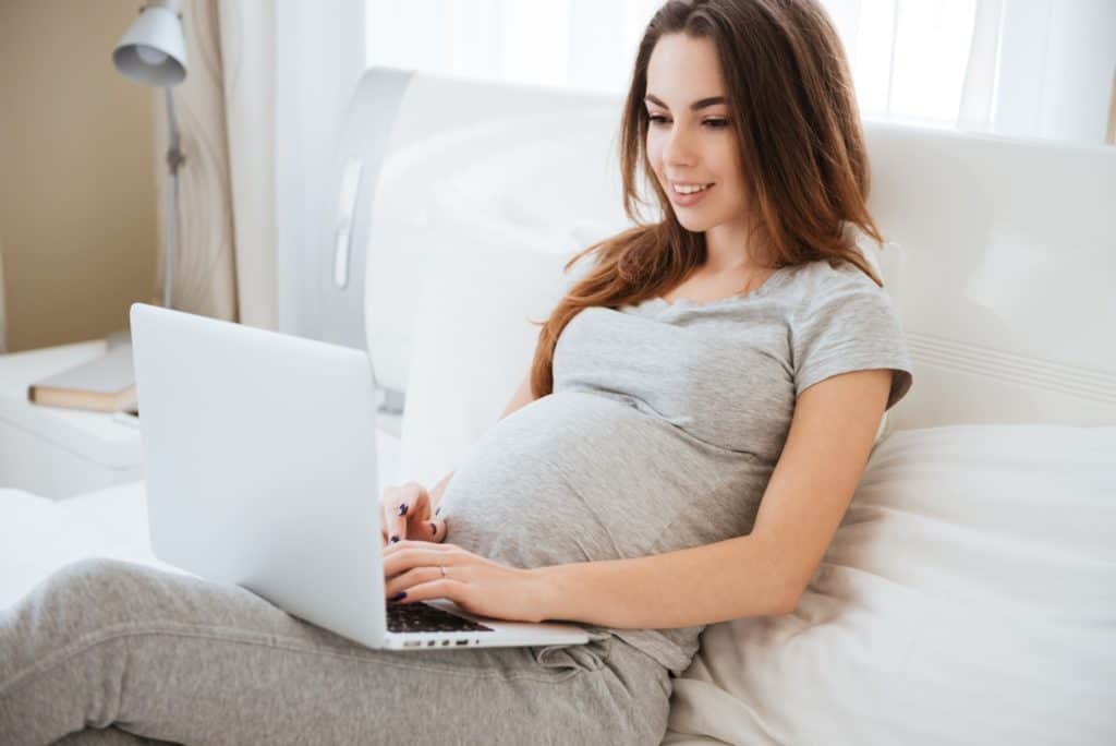 Cheerful pregnant young woman lying and using laptop on bed