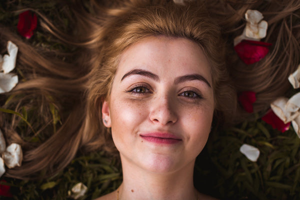 A girl with glowing skin smiling and laying down amongst rose petals. 
