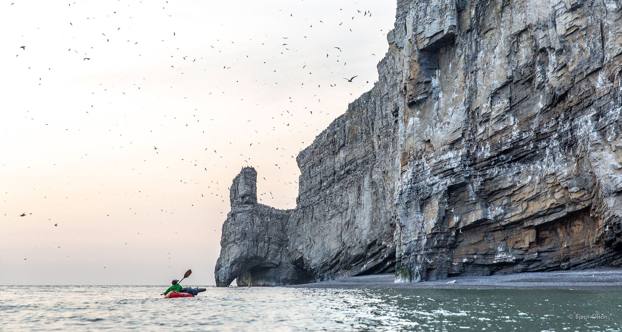 Ultralight packrafter looks at a swarm of birds over seaside cliff