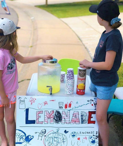 Two girls at a homemade lemonade stand on the sidewalk