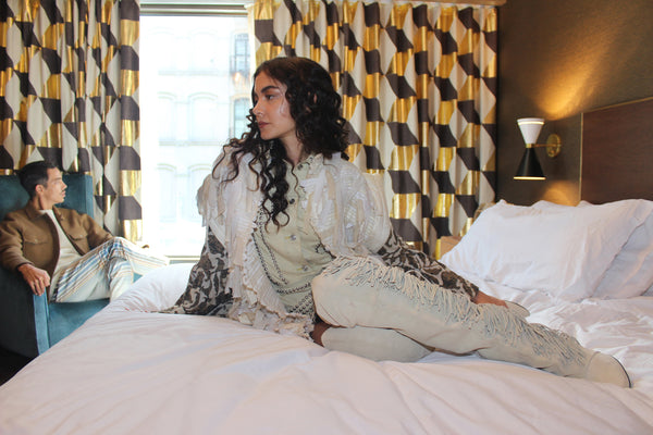 models posing in room, one on bed in foreground one on chair in front of window in chair.