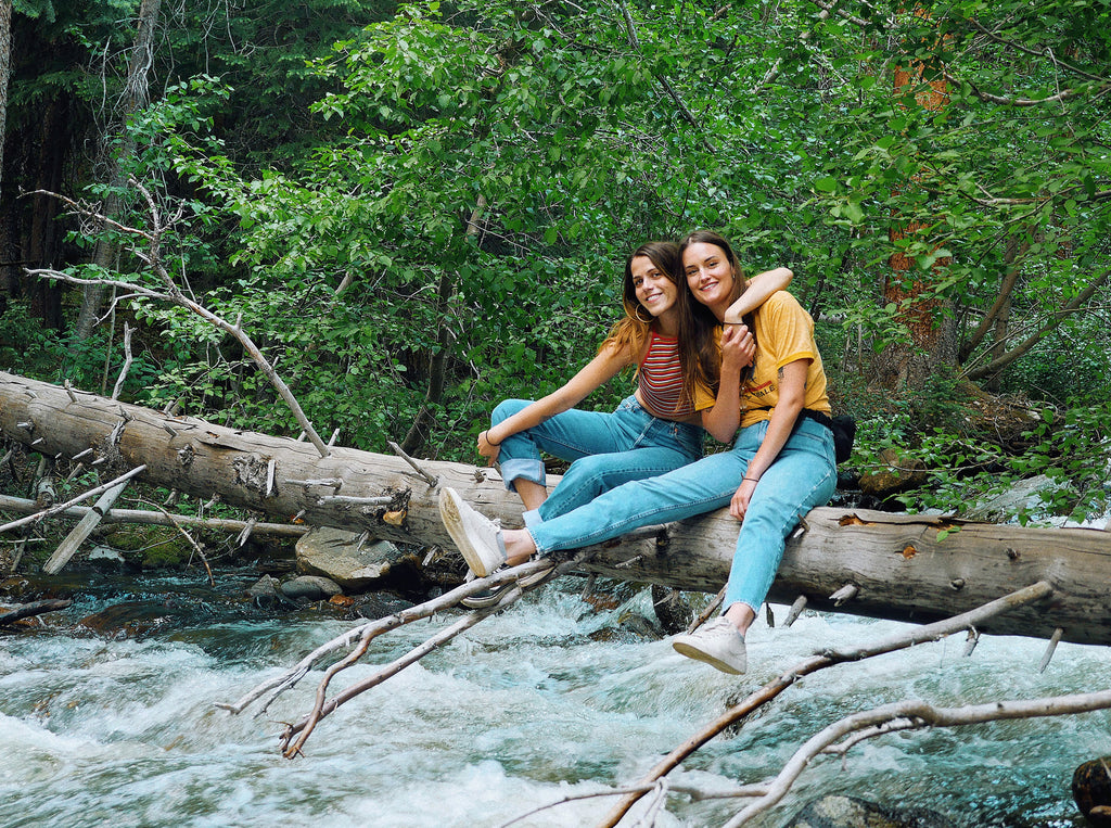 two people sitting on a fallen tree in a forest with river.
