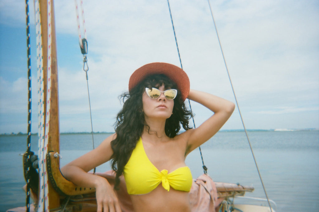 model in hat, sunglasses and a bikini posing on a sailboat with hand on head.