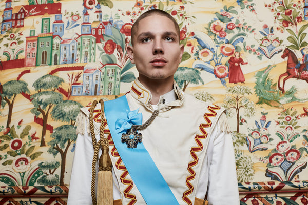 model in front of ornate wallpaper in ceremonial regalia and accoutrements.