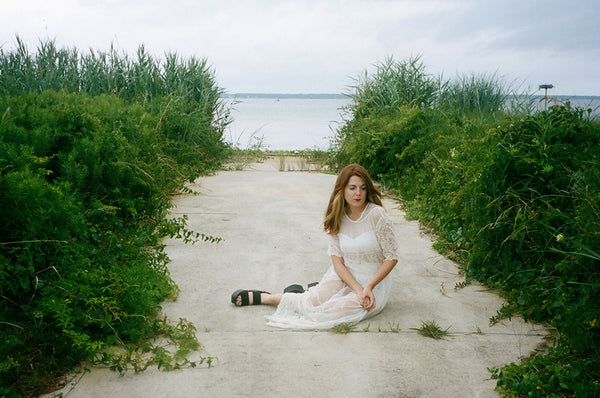 a person in a white dress sitting on a path near the water.