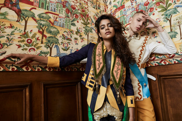 two models in corner of room in model in front of ornate wallpaper in ceremonial regalia and accoutrements.