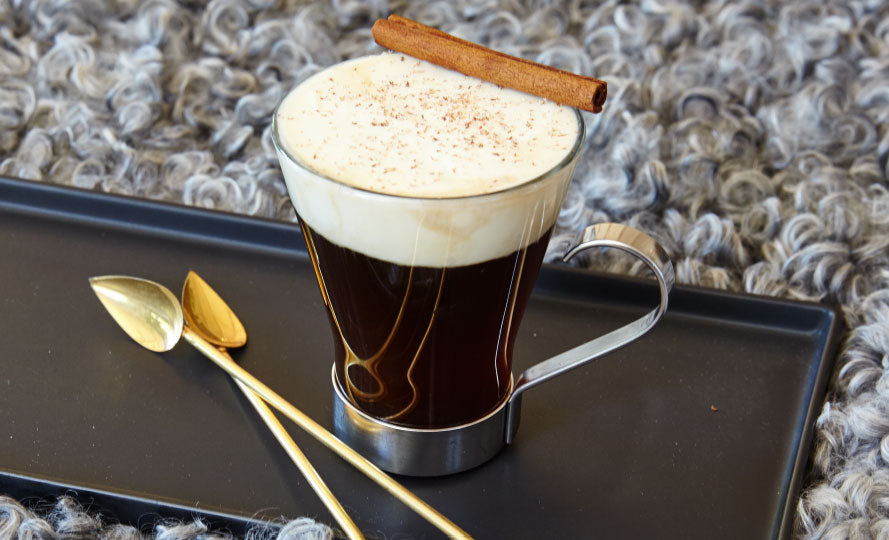 irish coffee in a clear glass with a large head of cream and a cinnamon stick resting on the edge