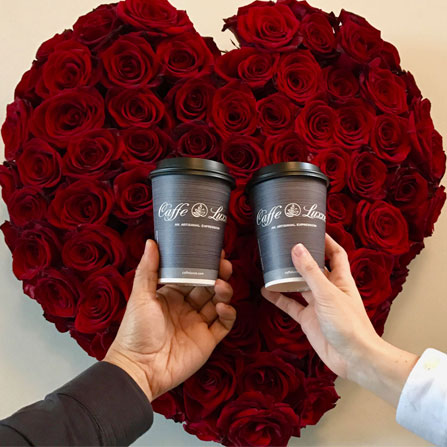 two people holding caffe luxxe coffee cups in front of a heart rose bouquet