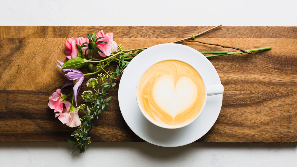 a latte with heart latte art sitting atop a wooden board with flowers directly above the latte
