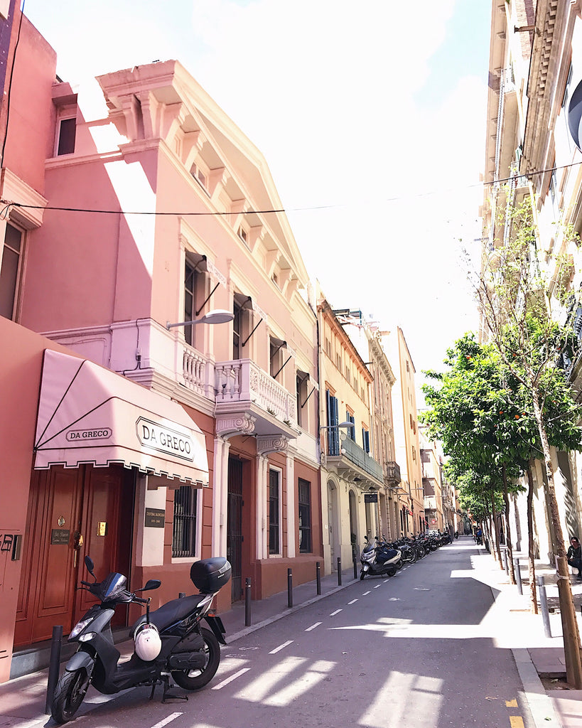 Street shot of Barcelona Spain, showing pink colored store fronts and a moped, on an empty street on a sunny day.