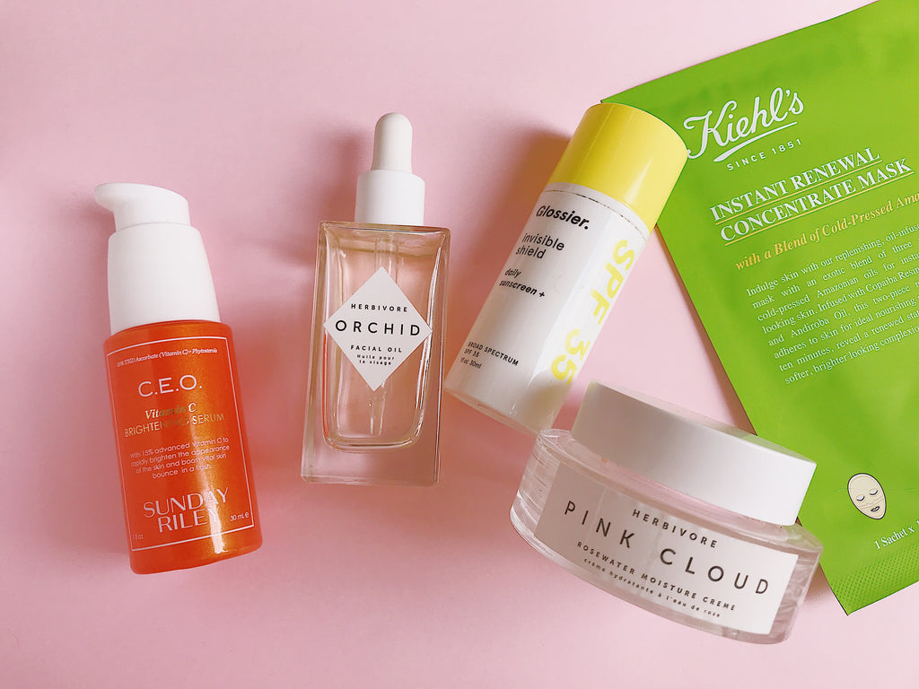 Skincare products against a pink background: C.E.O 15% Vitamin C Brightening Serum, Orchid Oil by Herbivore Botanicals, Pink Cloud Face Cream, glossier sunscreen and Kiehl's face mask.