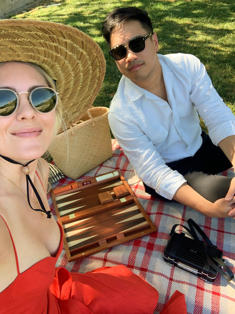 Katie Dean wearing a wide brim woven hat and red dress sitting on a checkered picnic blanket next to her husband with a wooden backgammon board game between them.