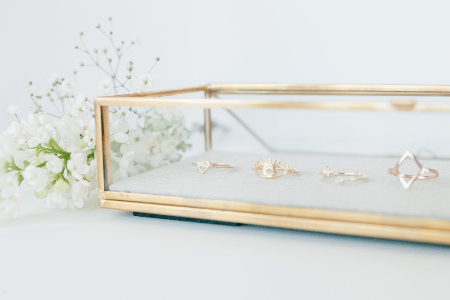 A gold-rimmed, glass display case with Katie Dean Jewelry rings displayed inside with white flowers next to the display.