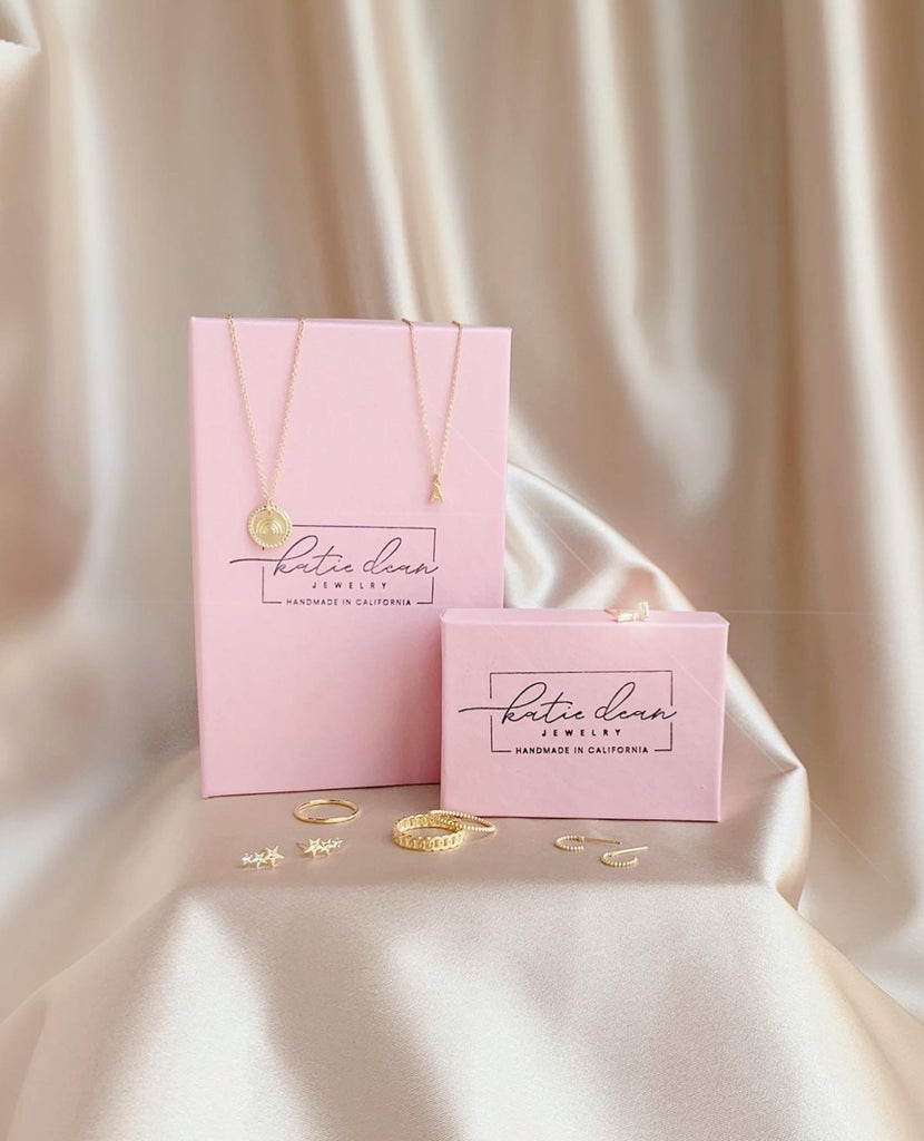 Katie Dean Jewelry pink boxes with hand stamped logo on top and dainty gold jewels around the boxes