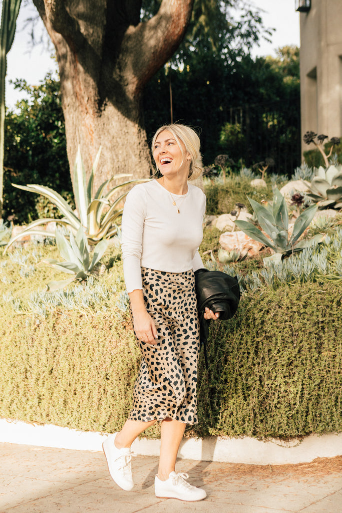 Katie Dean, founder of Katie Dean Jewelry, in a leopard print skirt, white tennis shoes and a white long sleeve shirt smiling and wearing her dainty gold jewelry while walking on a sidewalk in Los Angeles. 