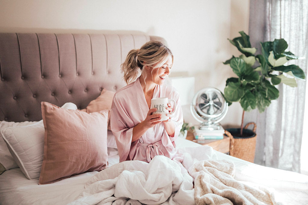Katie Dean sitting on a bed with a pink silk robe on and a white coffee mug, smiling with her blonde hair pulled back.