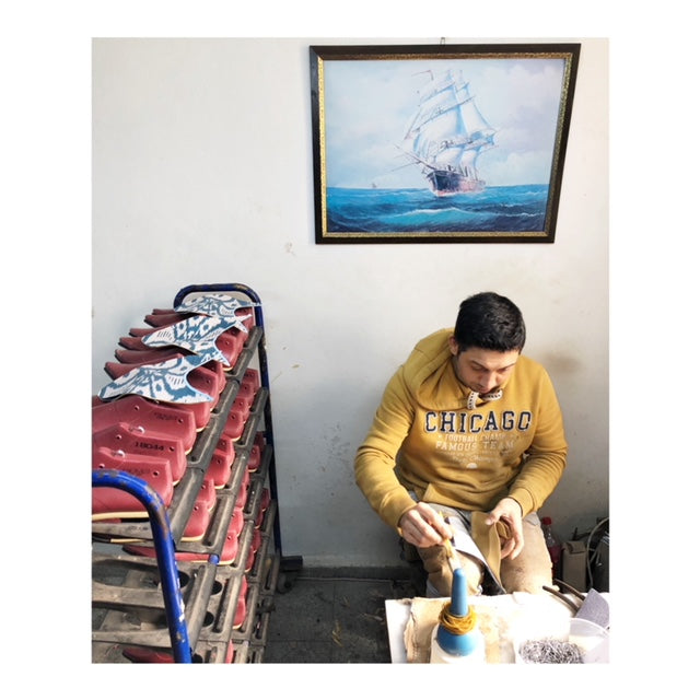 Cobbler in yellow working on kilim shoes and velvet slippers. He is seated in front of a white wall with a painting of a ship on it. 