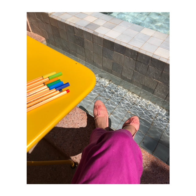 Sitting poolside in pink raffia loafers and pink pants, next to yellow table with colored drawing pens.