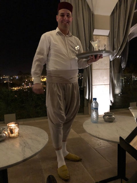 Waiter in Marrakech standing between two marble tables with drink on platter with view of Fez in background. 