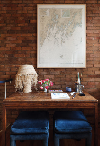 milicents brown working desk, covered with raffia lamp, paper, drawing utensils, and flowers. blue velvet chairs below. 