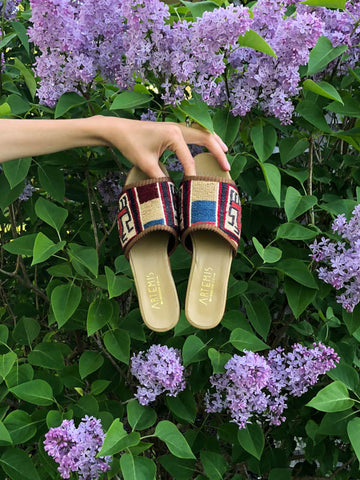 womens kilim sandals in sumak style being held in front of green plant with purple flowers. 