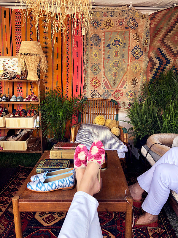 velvet slides resting on wooden table on kilim carpet surrounded by displayed kilim shoes, kilim loafers, and hung kilim carpets.