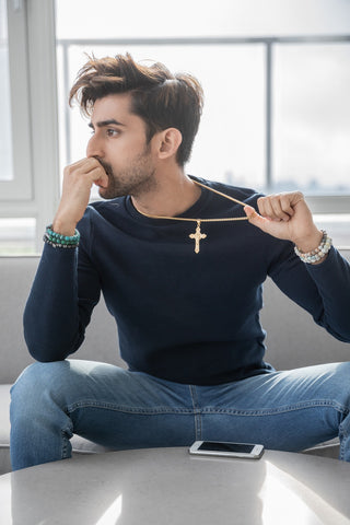 indian male model sitting on couch while modeling a gold stainless steel cross necklace and an assortment of men's natural stone bracelets 