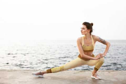 A woman doing yoga stretches by the ocean in leggings and a sports bra.