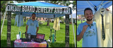 Drew smiling behind his booth at an outdoor venue. He has display cases in front of him filled with pendents, rings, cufflinks, earings and cards. There are two shirts in the background with Circuit Board Ties on them. A banner hangs above that reads "Circuit Board Jewelry and Art"