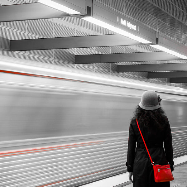 Back of woman standing at subway with small red purse, grey hat, and black long coat.