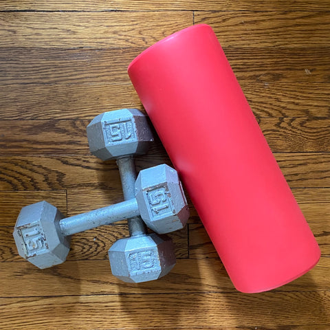 On a wood floor, seen from above, are two gray 15-lb dumbbells, one crossed over the other, and to their right a red foam roller lying on its side.
