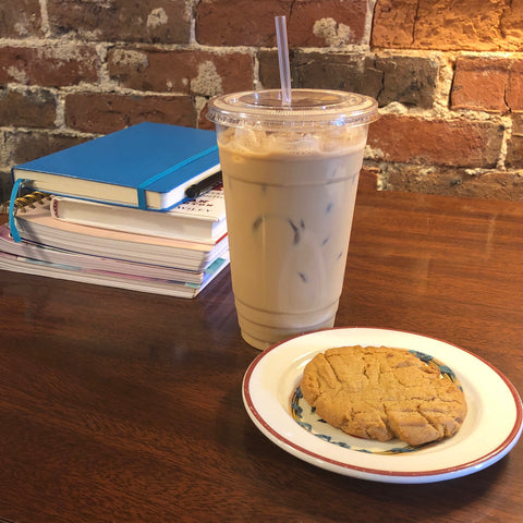 On a table sit a stack of books and notebooks, an iced latte, and a small plate with a cookie on it.