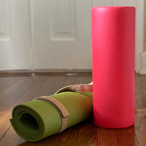 On a wood floor are a red foam roller, standing on end, and to its left a green yoga mat rolled in a tan strap.