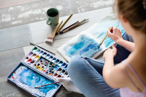 Woan sitting on floor with  watercolor paints, paper, coffee cup and art brushes on tthe floor. She is painting.