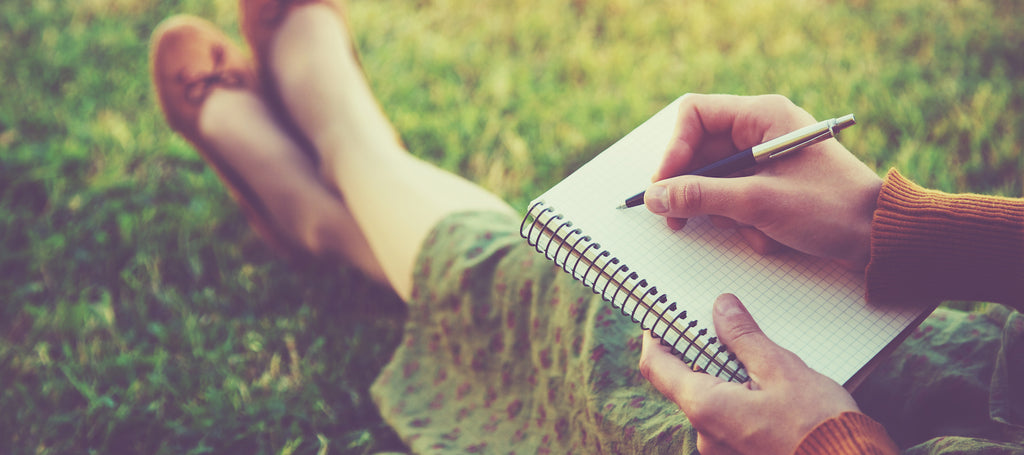 Woman sitting on grass with notebook on her lap, writing in it.