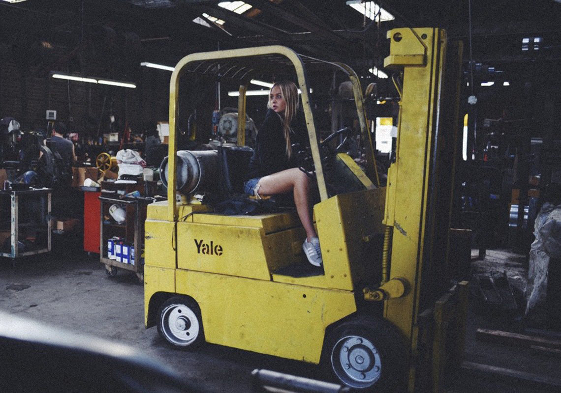 blond girl wearing black forrest and bob hoodie sitting on a yellow truck in a warehouse